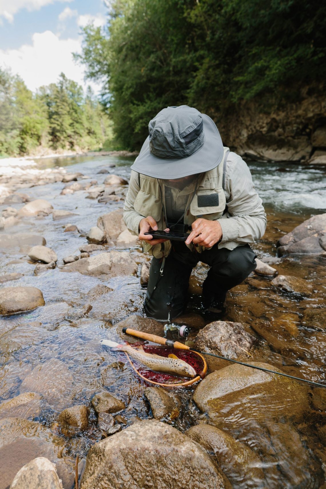 Fly fisherman photographing a trout in a landing net beside a rocky river